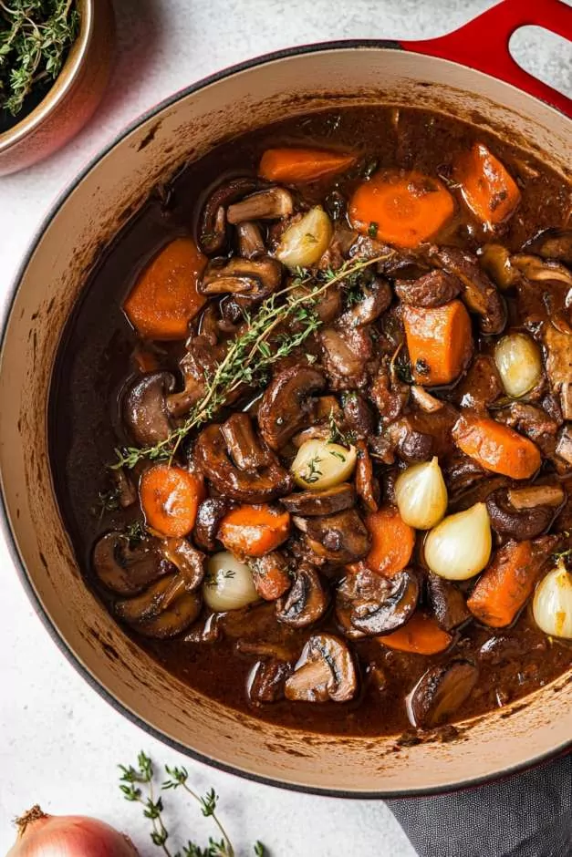 Hearty vegetable bourguignon in a red Dutch oven, featuring tender carrots, mushrooms, pearl onions, and herbs in a rich red wine sauce. Fresh thyme and bay leaves add depth to this savory, slow-cooked dish. A bowl of creamy mashed potatoes sits nearby, ready to be served.
