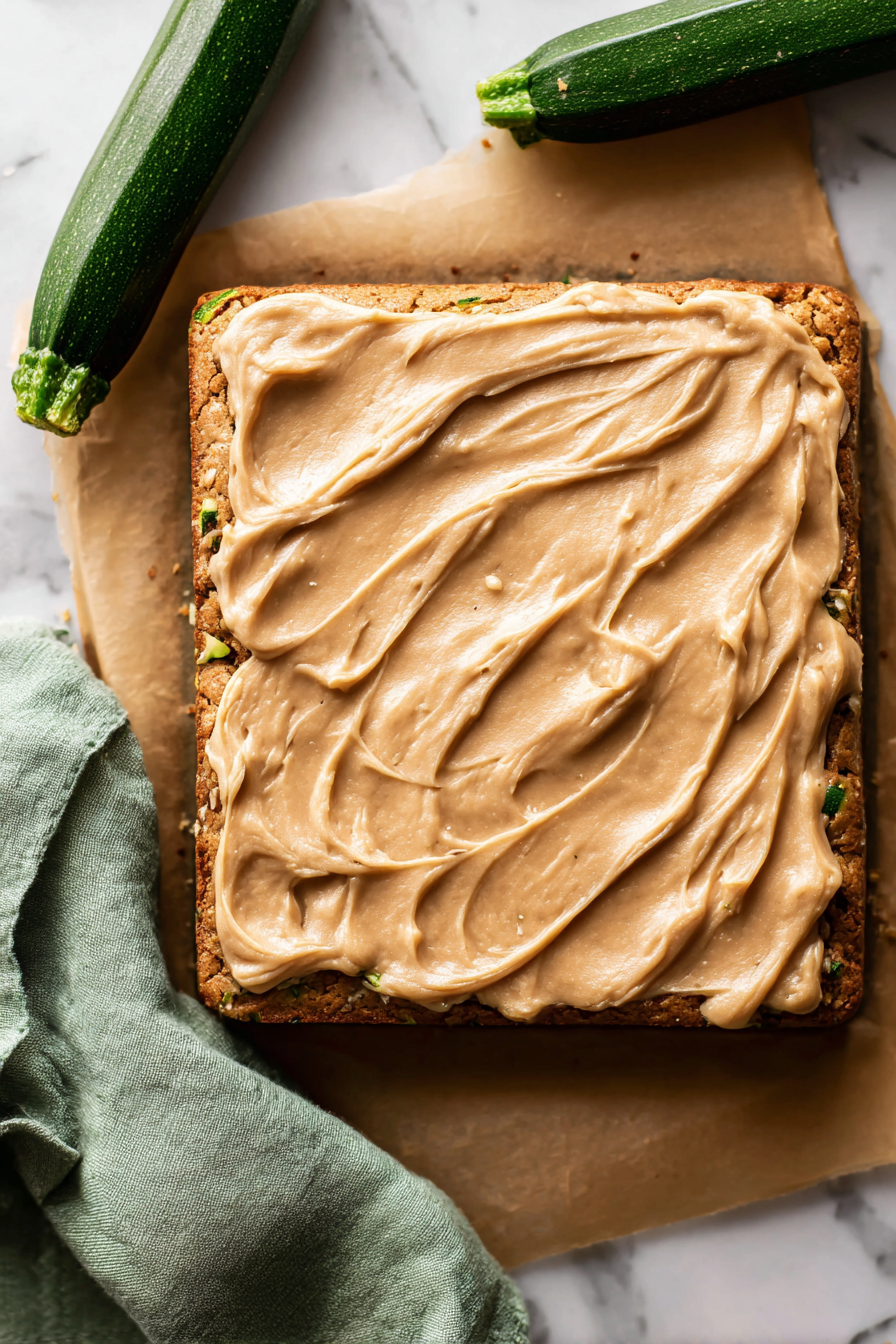a full uncut slab of zucchini pecan bars on light brown parchment paper, moist spiced zucchini cake base with visible chopped toasted pecans and subtle green zucchini flecks, topped with a thick smooth pale caramel-brown cream cheese frosting spread edge-to-edge