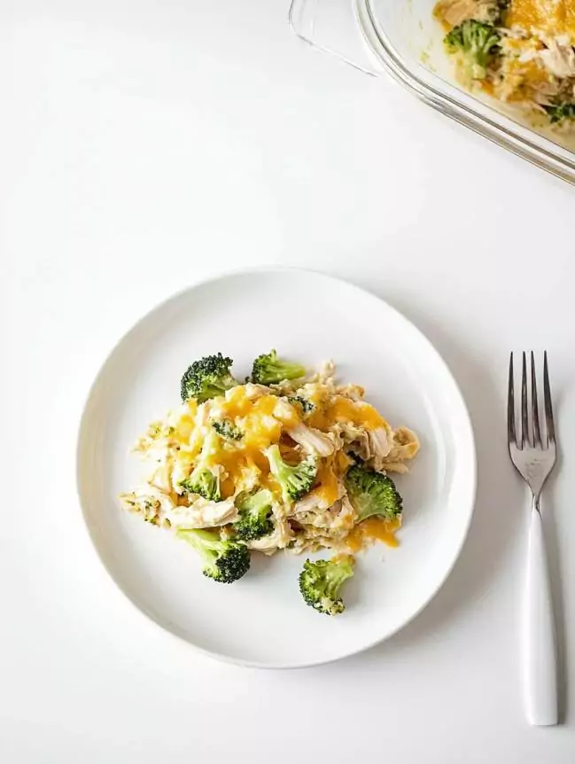 chicken broccoli casserole served on a white plate with a side of ranch dressing. This creamy, cheesy dish is made with shredded chicken, roasted broccoli, crispy bacon, and melted cheddar cheese, baked in a white casserole dish. A silver fork rests beside the plate. Perfect for a healthy, gluten-free dinner option.