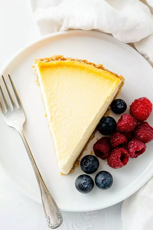 Slice of creamy cheesecake with a golden crust, topped with fresh raspberries, blueberries, and a drizzle of vibrant red berry sauce on a white plate. A delicious and visually appealing dessert.